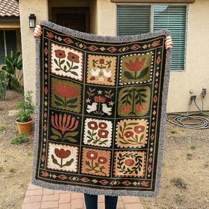 May include: A woven throw blanket with a black background and a repeating pattern of floral and bird motifs in shades of red, green, and beige. The blanket has a fringed edge.