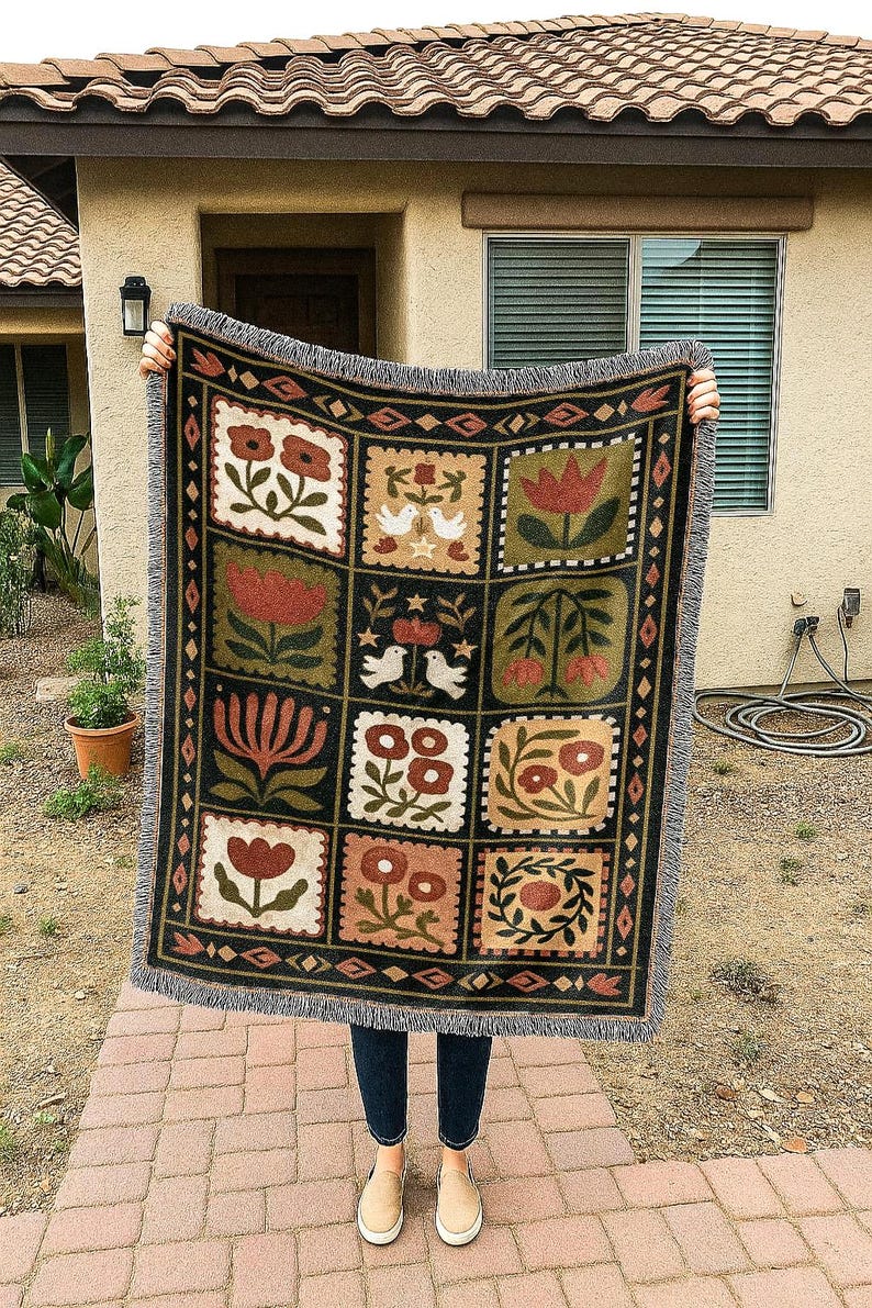 May include: A woven throw blanket with a black background and a repeating pattern of floral and bird motifs in shades of red, green, and beige. The blanket has a fringed edge.