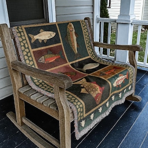 Puede incluir: Un sill&oacute;n mecedora de madera con una manta colorida con ilustraciones de peces. La manta tiene un borde verde oscuro y un borde con flecos. La silla est&aacute; en un porche con un exterior gris.