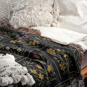 May include: A black floral throw blanket with a colorful fringe is draped over a bed. The blanket features a repeating pattern of yellow, orange, and purple flowers. Two white macrame pillows and white sheets are also visible.