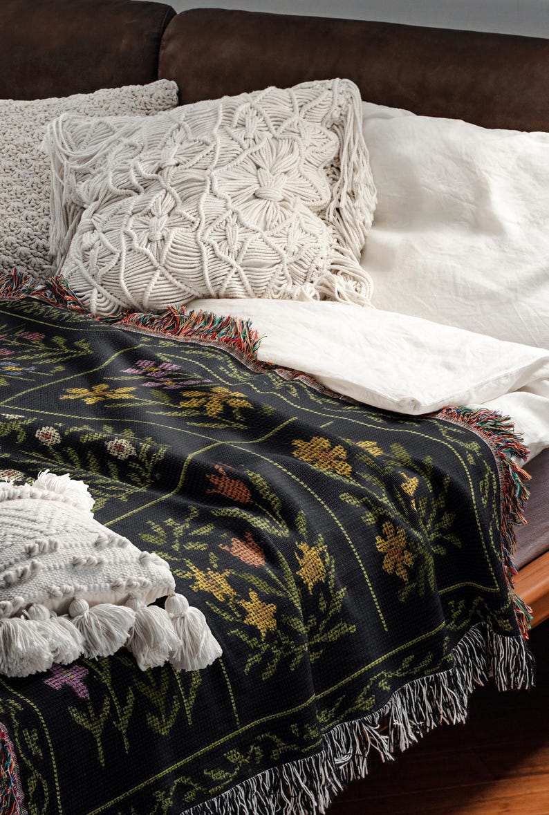 May include: A black floral throw blanket with a colorful fringe is draped over a bed. The blanket features a repeating pattern of yellow, orange, and purple flowers. Two white macrame pillows and white sheets are also visible.
