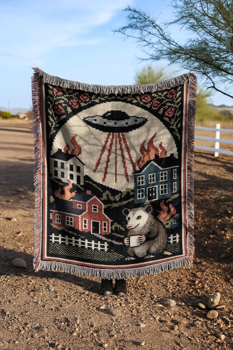 May include: Woven throw blanket with a black border and fringe. The design features a UFO, burning houses, and an opossum holding a mug. The top border has a floral pattern. The background is a light beige color.
