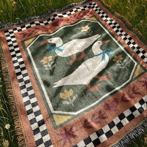 May include: A woven picnic blanket featuring a central design of two white geese with blue bows, surrounded by floral and leaf motifs on a green background. The blanket has a brown border with a black and white checkered pattern.