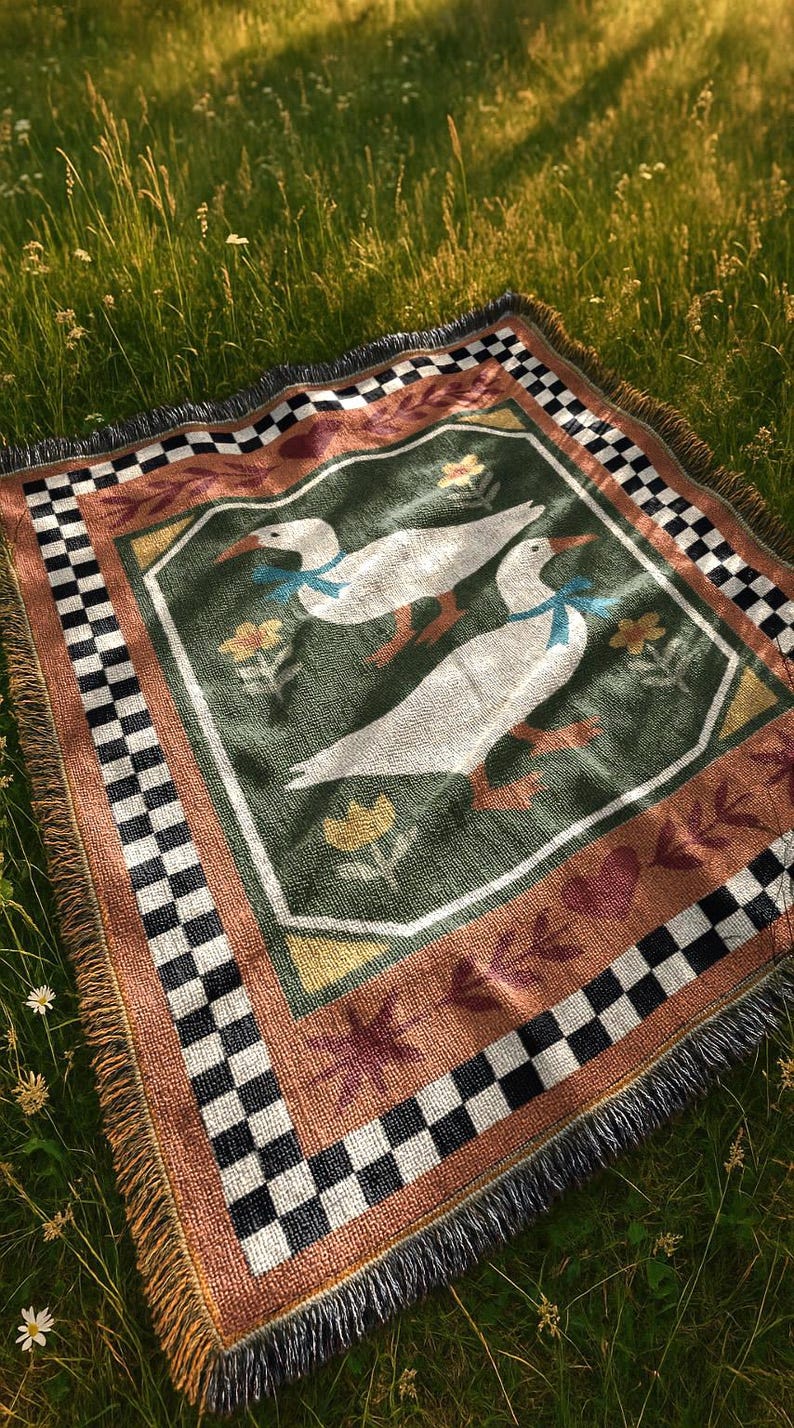 May include: A woven picnic blanket featuring a central design of two white geese with blue bows, surrounded by floral and leaf motifs on a green background. The blanket has a brown border with a black and white checkered pattern.
