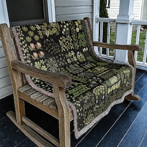May include: A wooden rocking chair with a botanical-patterned throw blanket. The blanket has a dark background with green and pink floral and leaf designs. The chair is made of light brown wood and sits on a porch.