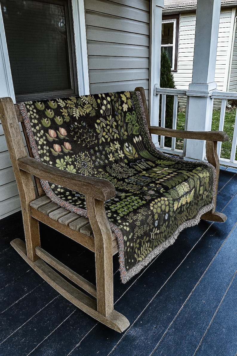 May include: A wooden rocking chair with a botanical-patterned throw blanket. The blanket has a dark background with green and pink floral and leaf designs. The chair is made of light brown wood and sits on a porch.