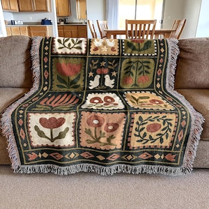 May include: A woven throw blanket with a patchwork design featuring floral and bird motifs in shades of green, brown, and cream. The blanket has a fringed edge and is draped over a brown sofa.