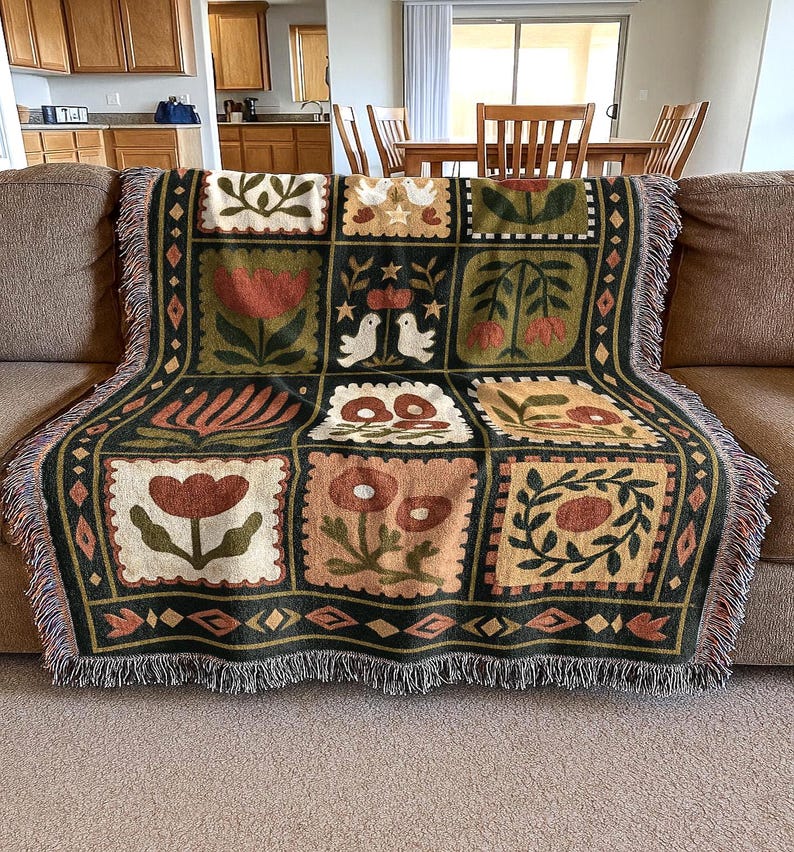 May include: A woven throw blanket with a patchwork design featuring floral and bird motifs in shades of green, brown, and cream. The blanket has a fringed edge and is draped over a brown sofa.