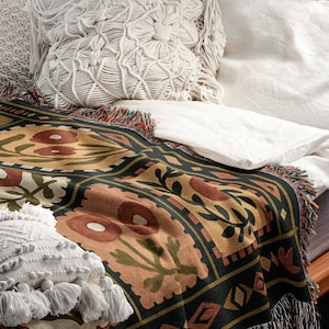 May include: A decorative throw blanket with a floral pattern in shades of brown, green, and gold, fringed edges, and a black border. Two white macrame pillows and a white sheet are also visible.