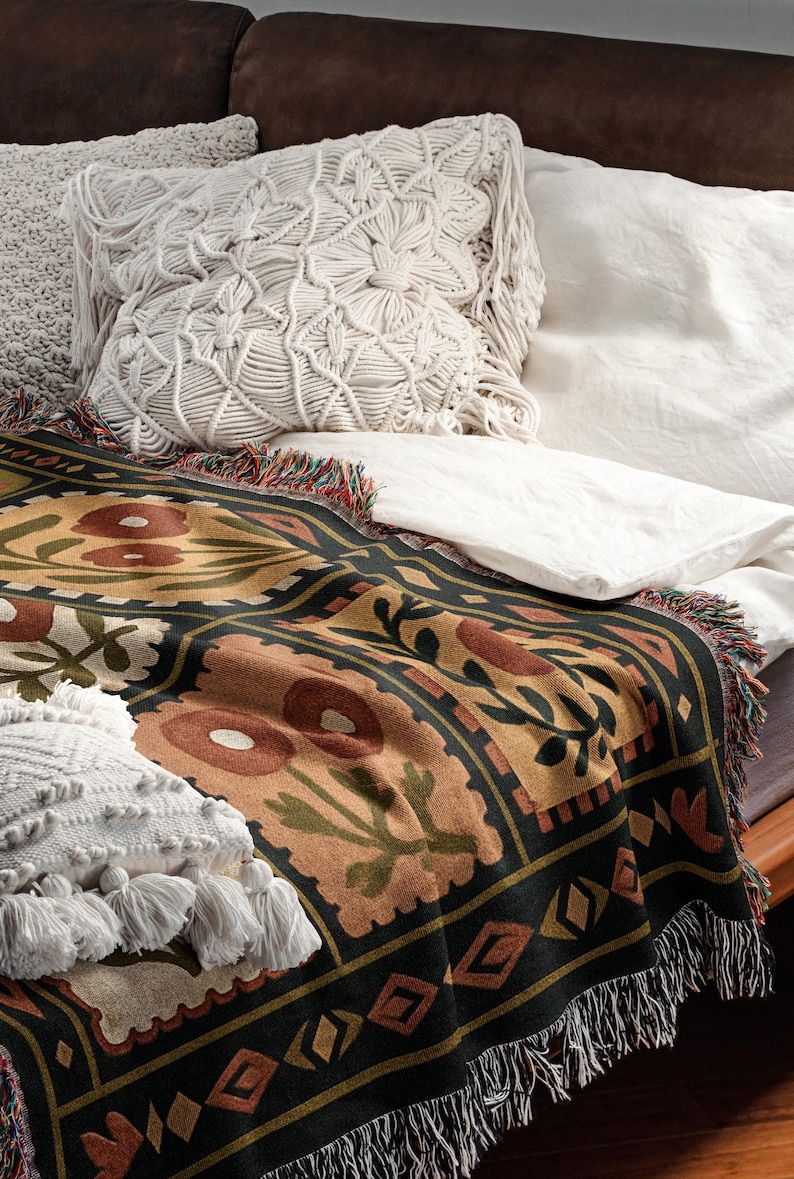 May include: A decorative throw blanket with a floral pattern in shades of brown, green, and gold, fringed edges, and a black border. Two white macrame pillows and a white sheet are also visible.