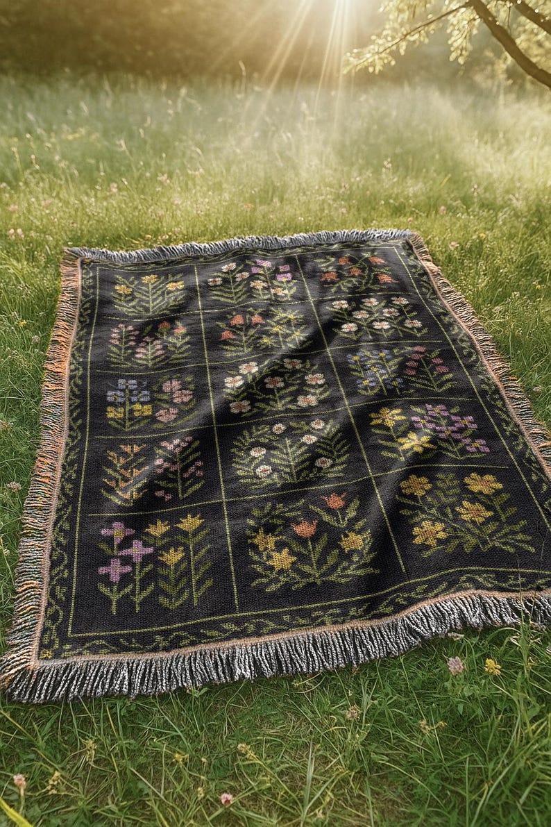May include: A black woven blanket with a floral pattern in various colors, including pink, yellow, and purple. The blanket is rectangular with a fringed edge and is laid out on a grassy surface.