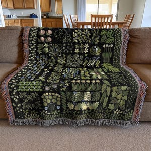 May include: A black throw blanket featuring a botanical design with green and blue foliage and flowers. The blanket, with a fringed edge, is draped over a brown sofa. The pattern showcases diverse plant illustrations.