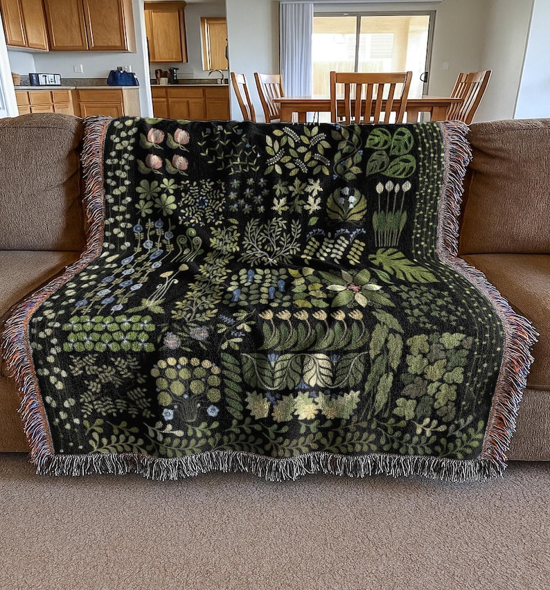 May include: A black throw blanket featuring a botanical design with green and blue foliage and flowers. The blanket, with a fringed edge, is draped over a brown sofa. The pattern showcases diverse plant illustrations.