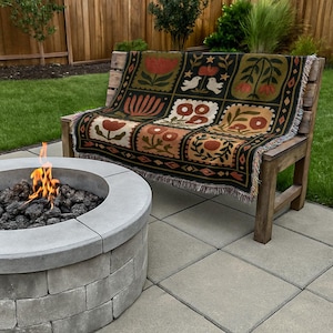 May include: A wooden bench with a decorative throw blanket featuring a floral pattern in shades of red, brown, and green. The blanket is draped over the bench, which is set on a patio with a fire pit in the foreground.