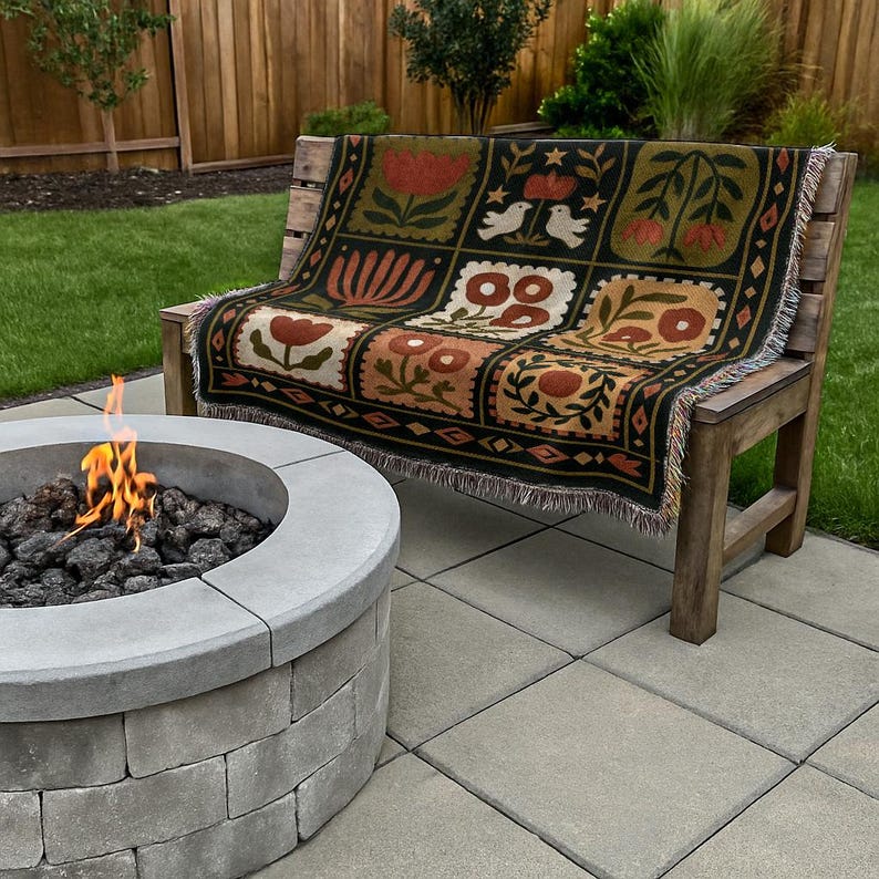 May include: A wooden bench with a decorative throw blanket featuring a floral pattern in shades of red, brown, and green. The blanket is draped over the bench, which is set on a patio with a fire pit in the foreground.