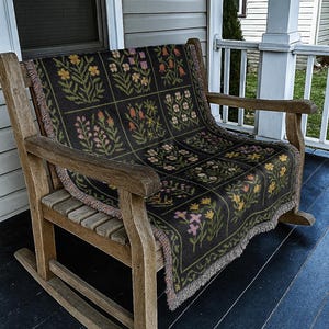 May include: A wooden rocking chair with a black floral patterned throw blanket. The blanket features a grid of colorful flowers in pink, yellow, orange, and green. The chair is on a dark blue porch.