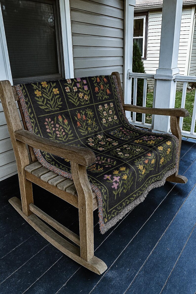May include: A wooden rocking chair with a black floral patterned throw blanket. The blanket features a grid of colorful flowers in pink, yellow, orange, and green. The chair is on a dark blue porch.