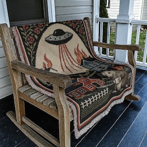 May include: A woven throw blanket with a whimsical design featuring a UFO, flames, houses, and a possum holding a mug. The blanket is draped over a wooden rocking chair on a porch. The color palette includes beige, black, red, and green.
