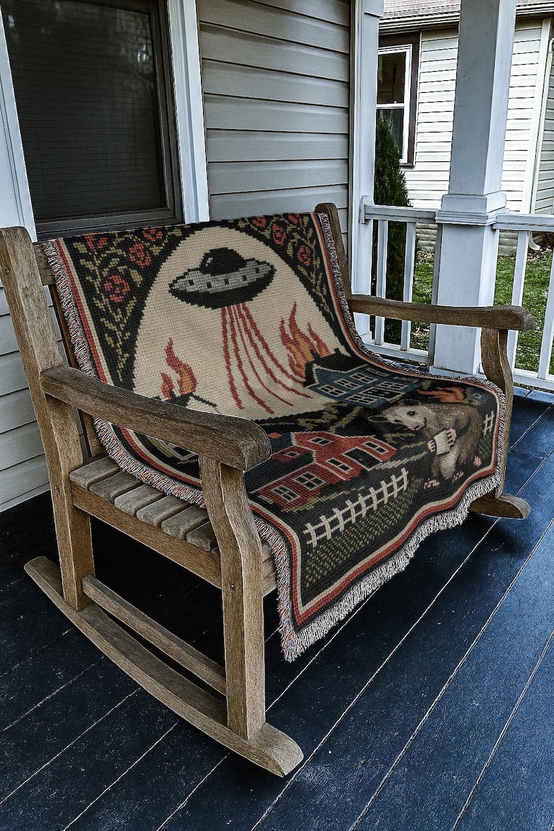 May include: A woven throw blanket with a whimsical design featuring a UFO, flames, houses, and a possum holding a mug. The blanket is draped over a wooden rocking chair on a porch. The color palette includes beige, black, red, and green.