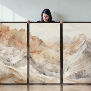 May include: A triptych of framed art prints depicting a watercolour mountain landscape in neutral tones of beige, brown, and grey. The artwork is displayed in a room with a wooden floor and a plant.