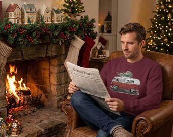 Peut inclure: Un sweat-shirt à manches longues bordeaux avec une impression graphique d'un van vintage rouge et blanc transportant un sapin de Noël sur son toit. Le sweat-shirt a un col rond et est porté avec un jean bleu foncé et des chaussettes beiges.