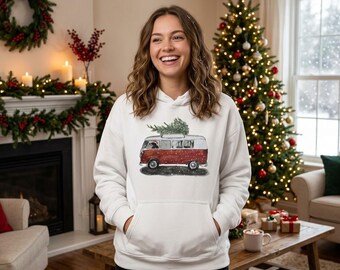 Peut inclure: Un sweat-shirt à capuche blanc à manches longues avec une impression graphique représentant un van de style vintage rouge et blanc transportant un petit sapin vert sur son toit. L'illustration du van inclut des fenêtres sombres et de la neige.