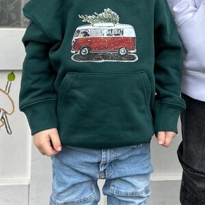 Op de afbeelding: Een donkergroene hoodie met een grafische print van een rood-witte vintage busje met een kerstboom op het dak. Het ontwerp van het busje bevat sneeuwdetails en bevindt zich boven een kangoeroezak aan de voorzijde van het kledingstuk.