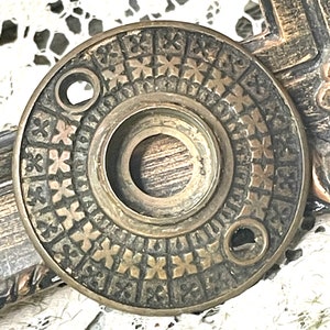 May include: Antique brass door knob backplate with a detailed, embossed floral pattern. The circular plate features a central opening and two screw holes. The aged metal has a warm, vintage aesthetic, suggesting a classic or vintage style.