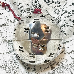 May include: A spherical silver-toned metal container featuring a series of cut-out heart and geometric shapes across its surface. The object includes a central vertical latch mechanism and a dark red braided cord attached to the top for hanging.