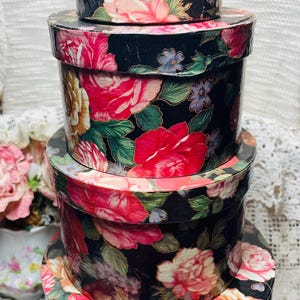 May include: A stack of four round decorative boxes. The boxes are black with a floral pattern featuring pink, red, and yellow roses, blue flowers, and green leaves. The boxes are of varying sizes, with the largest at the bottom.