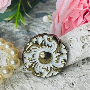 May include: A round, antique-style button with a white and gold floral design. The button has a raised center and ornate detailing. The button is set against a backdrop of lace, pearls, and pink flowers.