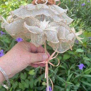 May include: A decorative miniature umbrella crafted from cream-colored lace with multiple ruffled tiers. The handle and canopy are accented with peach-colored satin ribbons, small fabric flowers, and a delicate ring of white faux pearls at the top.