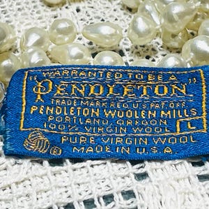 May include: A blue Pendleton label with gold lettering, featuring text like "Pendleton Woolen Mills" and "Pure Virgin Wool." The label is on a white lace surface, with a strand of off-white pearls in the background.