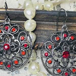 May include: A pair of black filigree earrings with red rhinestone accents. The earrings are round and have a delicate, intricate design.