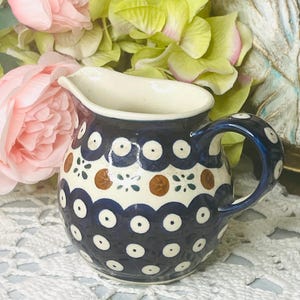 May include: A small, ceramic creamer with a dark blue base and white and brown polka dot pattern. The handle and rim are also dark blue. The creamer is decorated with a floral design and has a white interior.