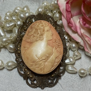 May include: Oval cameo brooch featuring a woman's profile in relief, set in an ornate bronze-toned frame. The cameo is a light peach color, and the frame has intricate filigree details. A pearl necklace and a pink rose are in the background.