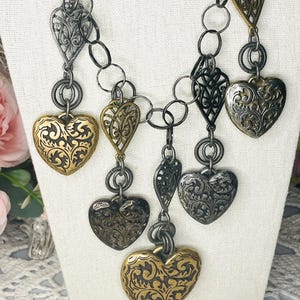 May include: A multi-strand necklace featuring various heart-shaped charms in gold and silver tones. The charms display intricate embossed scrollwork and openwork filigree patterns, suspended from dark metal circular links and a matching chain.