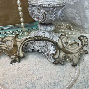 May include: A decorative drawer pull featuring an ornate, scrolled design with a distressed finish in shades of cream, beige, and dark brown. The hardware has a curved, elongated shape with intricate relief detailing and symmetrical flourishes.