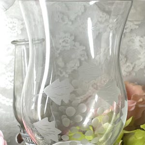 May include: Clear glass vase with a wide, curved shape, resting on a white base. The vase features an etched design of grapes and leaves. The background includes a glimpse of a vintage oil lamp and floral arrangements.