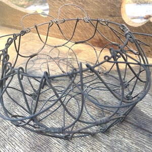 May include: A handcrafted, dark gray wire basket with a circular design. The basket features a series of interconnected wire loops and a handle. The basket is made of metal and is suitable for decorative or functional use.