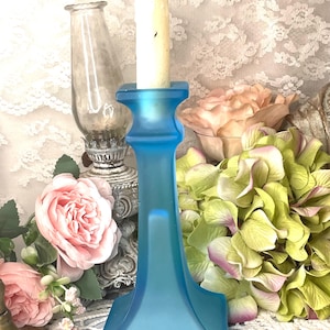 May include: A light blue frosted glass candlestick holder with a lit white candle. The holder has a square base and a flared design. The background includes a vintage oil lamp, pink roses, and green hydrangeas.