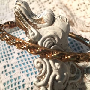 May include: A gold-colored bangle bracelet with a detailed, textured design. The bracelet is resting on a white and gold decorative object, with a lace background. The bracelet's intricate pattern adds a touch of elegance.