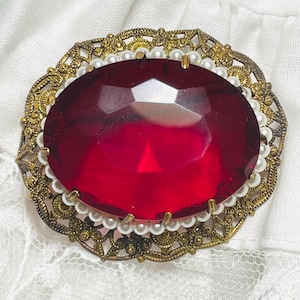 May include: An oval brooch featuring a large, faceted deep red stone set in the center. The stone is surrounded by a ring of small white pearls. The outer frame consists of an intricate, openwork gold-tone metal filigree design.