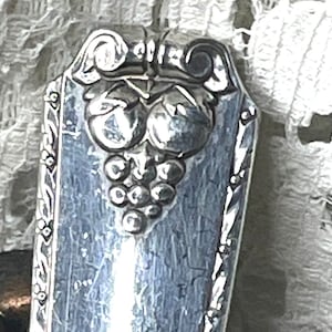 May include: Close-up of a silver-toned serving utensil, possibly a spoon or fork, with a decorative grape cluster design. The handle has an ornate border and a rectangular shape. The utensil is set against a textured, off-white background.