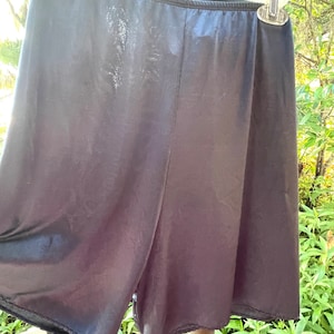 May include: A pair of solid black shorts with a smooth, slightly reflective finish. The shorts feature an elasticized waistband and are suspended from a clear plastic hanger, showcasing a simple, unadorned design with clean, straight hemmed edges.