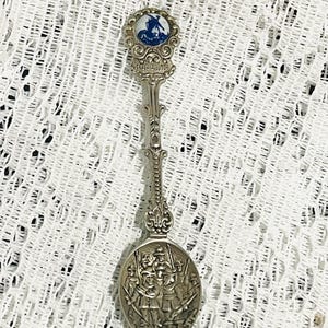 May include: A silver-toned souvenir spoon with a detailed design. The spoon features a small, round emblem with a windmill scene and the word "Holland". The bowl of the spoon depicts a group of figures. The spoon is approximately 5 inches long.