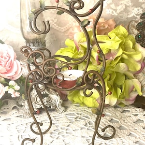 May include: A decorative metal candle holder with a scrolled design, painted in a bronze color. The candle holder features green leaves and red berry accents. A lit white candle sits in the center, adding a warm glow. The overall design is ornate and vintage.