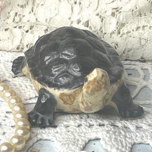 May include: A ceramic turtle figurine with a dark gray shell and tan and black details. The turtle is posed on a white lace surface, with a strand of faux pearls in the foreground. The figurine is a decorative item.