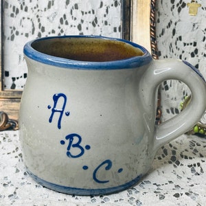 May include: A handmade stoneware mug with a light gray body, a blue rim, and a blue handle. The mug features the letters "A", "B", and "C" in blue script. The interior of the mug is a warm brown color.