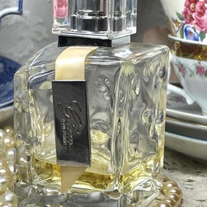 May include: A clear, square perfume bottle with a clear, rectangular cap. The bottle has a gold ribbon and a black label with the word "Glam" in silver. A strand of pearls and floral teacups are in the background.